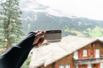 Naklejka premium Hand holds a small cup of coffee overlooking Grindewald, Switzerland in the morning