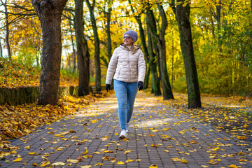 Beautiful middle-aged blonde woman wearing jeans, beige jacket and winter hat and sunglasses walking on alley full of yellow fallen leaves in park on autumn day. Front view. Autumnal walk in city park