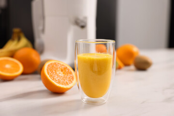 Glass of fresh juice, modern juicer and fruits on white marble table in kitchen, selective focus