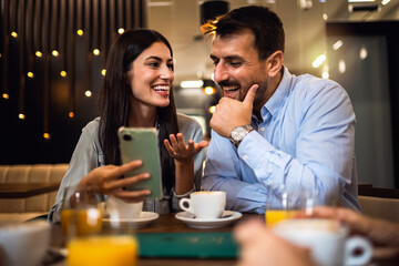 Friends met at the cafe. Drinking coffee and orange juice. The guy is showing something on his phone to his girl