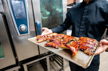 A chef in a professional kitchen presents a tray of glazed ribs, showcasing the delicious and visually appealing dish. Ideal for culinary, restaurant, or food-related content.
