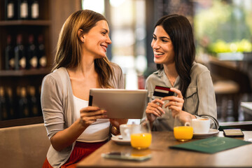 Two girl best friends met at the cafe to shop online together. They are using tablet and credit card. Friends hang out and drink coffee and orange juice.