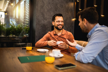 Two male friends met at the cafe to hang out. They are laughing, having a good time and they drink orange juice and coffee.