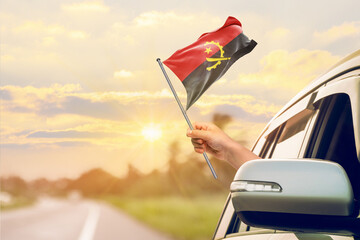 Waving the Angola flag against the sunrise or sunset from a car driving along a country road. Holding the Angola flag, traveling by car, on a weekend trip.