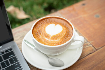 Close-up of Hot coffee latte with latte art milk foam in cup mug and laptop computer on wood desk office desk in coffee shop at the cafe,during business work concept