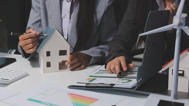 Diverse team with Asian man, African American woman, Caucasian woman discussing a clean energy city planning project with building models and solar panels.