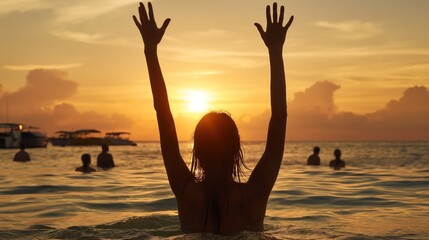 Fototapeta premium A silhouette of an individual raises their arms in joy as the sun sets over the ocean, reflecting golden hues on the water with others swimming nearby