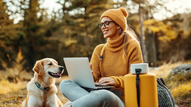 Woman wearing a yellow sweater and beanie, sitting outdoors with a laptop on her lap and a golden retriever dog beside her.