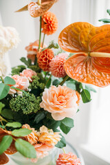  Close-up of a vibrant floral arrangement featuring a striking orange anthurium surrounded by soft peach roses and green foliage.