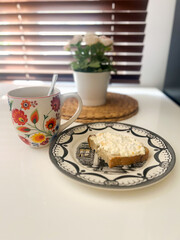 A cozy breakfast setting featuring a colorful floral mug of coffee and a plate with a cottage cheese toast. A peaceful morning vibe with a flower pot in the background.