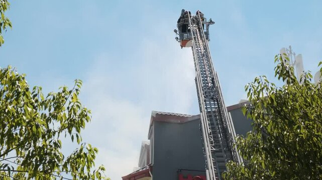 Back view of firefighters in elevated platform extinguish burning house using extended ladder equipped with water hose to spray water onto the building roof with smoke. Fireman extinguishing fire