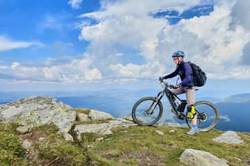 Fototapeta premium Cyclist man riding electric bike outdoors. Male tourist resting on the top of hill, enjoying beautiful mountain landscape, wearing helmet and backpack. Concept of active leisure.