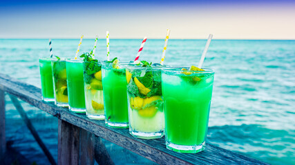 Sept verres à cocktails alignées sur une balustrade en bois, devant la mer et une lumière de soleil couchant
