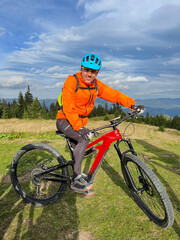 Obraz premium Cyclist man riding electric bike outdoors on sunny day. Portrait of smiling male tourist resting on grassy hill, enjoying beautiful mountain landscape, wearing helmet. Concept of active leisure.