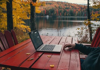 Man working on his laptop outdoors near a lake surrounded by autumn foliage. The red wooden furniture contrasts with the colors of nature, creating a cozy and calm atmosphere for remote work or study
