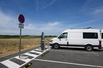 Side view of an electric van charging at a charge station in the middle of the fields. A sign indicates no parking.  A pictogram authorizes electric vehicles.
