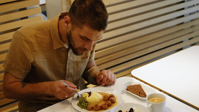 A young Caucasian bearded man has a delicious and satisfying dinner at an IKEA restaurant. Mashed potatoes with green peas, meat meatballs and gravy. Cappuccino coffee with cake for dessert