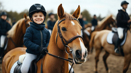 A smiling child wearing a helmet sits on a horse, surrounded by other riders in equestrian gear, during a group horseback riding activity.