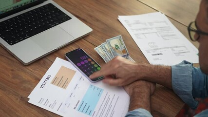 Middle aged accountant subtracts amount of dollars on table by phone calculator, man in glasses thinks over budget for month taking into account household expenses slow motion