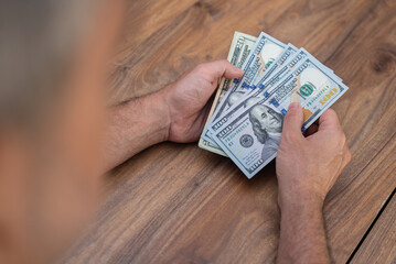 Person holding a stack of US dollar bills over a wooden table. Financial wealth and cash management concept. Ideal for topics related to personal finance, banking, and economic activities 