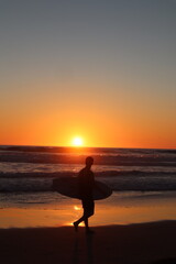 Fototapeta premium Surfer silhouette walking on the beach during orange sunset at Pacific Beach, San Diego California
