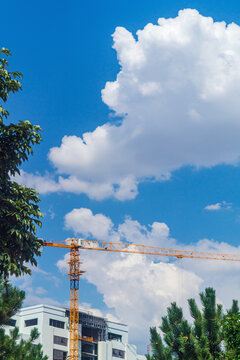 Blurred large white cumulus clouds fly across the sky behind yellow construction crane at a building site on a sunny summer day, foreshadowing possible construction stoppage due to changing weather