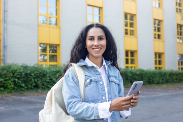 Half-length portrait of an Arab woman standing in front of an educational institution on a university campus holding a phone typing messages looking at the camera smiling brightly wearing a denim jack