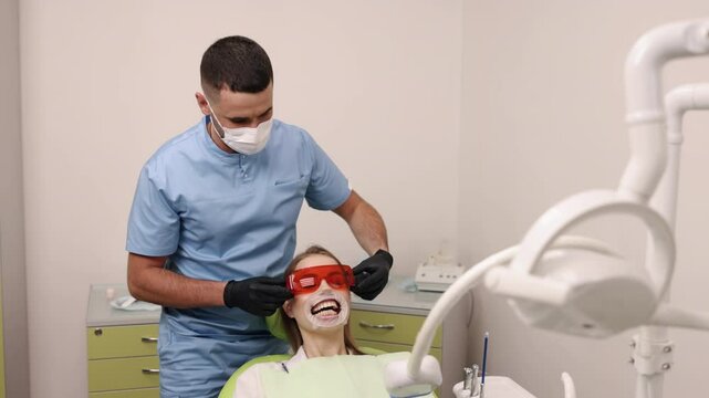 Mouth retractor, Dental tools, Dental visit. Dentist in blue scrubs and mask is adjusting protective eyewear on smiling patient who has mouth retractor in place while seated in dental chair.
