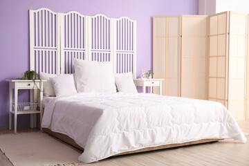 Interior of stylish bedroom with comfortable bed, white pillows, bedside tables and folding screen