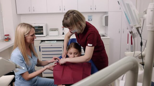 Red scrubs, Dental clinic, Clinic interior. Dental assistant in red scrubs is putting bib on young girl seated in dental chair while dentist in blue scrubs looks on.