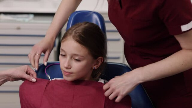 Dental chair, Oral care, Dental treatment. While dentist in blue scrubs watches, dental assistant in red scrubs puts bib on young girl sitting in dental chair.