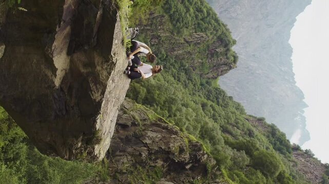 Two young girls decided to take a break from their hike and rest on the top of a steep cliff. They were surrounded by the green mountains with peaks of indescribable beauty. Slow motion. Vertical.