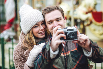 Beautiful young loving couple, boyfriend and girlfriend having fun on a Christmas market wearing warm clothes, taking pictures on old retro camera. Outdoors, winter time, snowy weather.