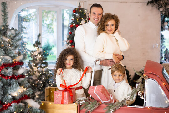 Mother and father with gift boxes in their hands and near are theis children in red car near Christmas trees.