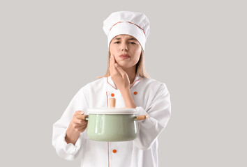 Young female chef with cooking pot suffering from tooth ache on grey background