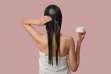 Beautiful young woman with jar of cosmetic product applying hair mask on pink background, back view