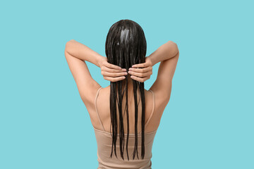 Beautiful young woman applying hair mask on blue background, back view