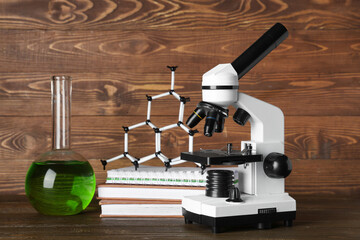 Modern microscope with chemical flask, books and molecular model on table against wooden background