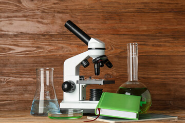 Modern microscope with chemical flasks, petri dish and book on table against wooden background
