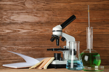 Modern microscope with chemical flasks, petri dish and book on table against wooden background