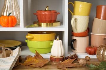 Shelf unit with dishware, pumpkins and autumn leaves near light wall