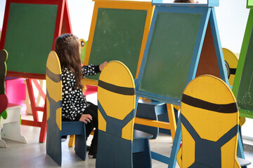 Little girl drawing with chalks on green blackboard in a kindergarten. Child in initial level...