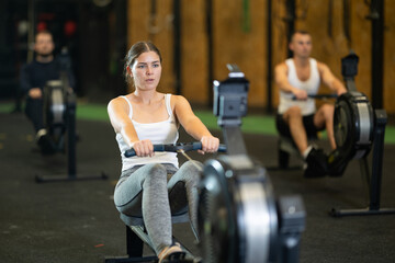 Determined young girl training with rowing machine in sports hall during crossfit workout