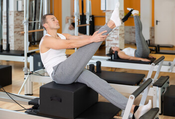 Concentrated athletic young adult man performing set of pilates exercises on reformer with sitting box during group workout in fitness studio. Healthy active lifestyle....