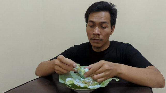 A Southeast asian man putu cake (traditional food indonesia)  with grated coconut and white sugar on a plate lined with banana leaves, sweet food, eating cake, 4K.
