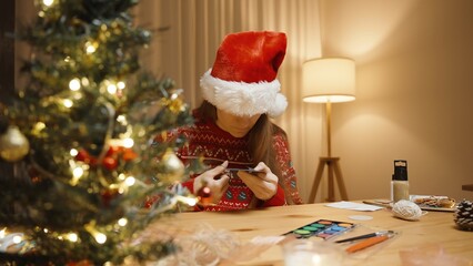 Woman Wearing a Santa Hat and Festive Sweater, Focusing on Crafting, Surrounded by Holiday Decorations and Warm Candlelight.