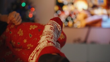 Person in holiday-themed pajamas and festive socks rests on a couch, with blurred Christmas lights and decorations creating a cozy, seasonal backdrop.