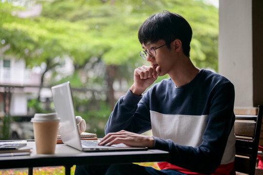 A thoughtful young Asian man is working remotely from an outdoor table, working on his laptop.