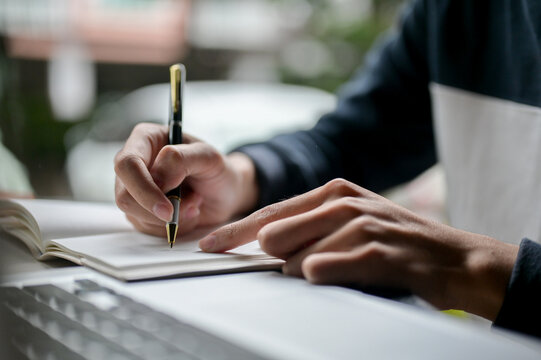 A male college student writing in his notebook or taking notes while studying online on his laptop.