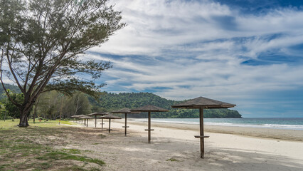 A row of sun umbrellas on the sandy beach. The waves of the turquoise ocean are foaming. A green hill against a background of blue sky and clouds. Malaysia. Borneo. Kota Kinabalu.